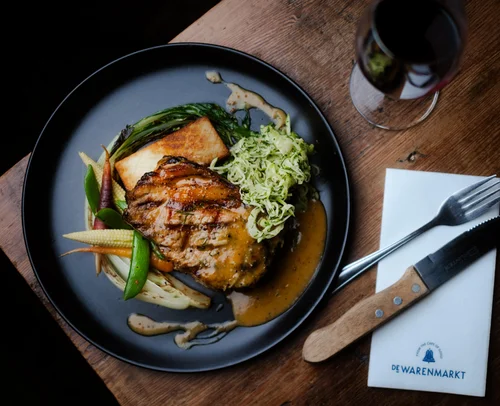Gourmet pork dish with vegetables and sauce, served on a black plate with wine.